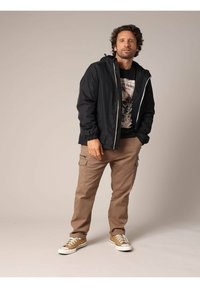 Zwarte lichtgewicht jas met capuchon, ritsluiting en elastische manchetten, gecombineerd met beige cargobroek en tan sneakers. Grafisch t-shirt eronder.