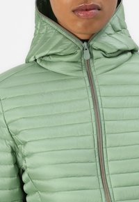 Lys grøn pufferjakke med hætte, der har vandret quilting, en frontlynlås og en glat, skinnende overflade.