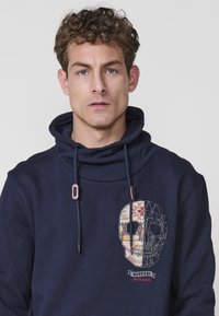 Sudadera con capucha azul marino que presenta un gráfico de calavera en el frente con patrones y líneas intrincadas, tejido texturizado y cordones ajustables.