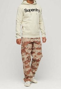 Man wearing beige Superdry hoodie, brown and tan camouflage cargo pants, and white sneakers standing against plain background.