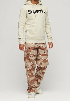 Man wearing beige Superdry hoodie, brown and tan camouflage cargo pants, and white sneakers standing against plain background.