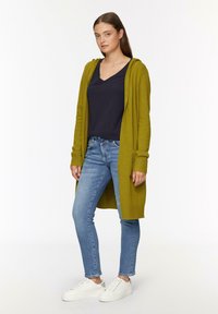 Lang olijfgroene cardigan met capuchon, gecombineerd met een marineblauwe V-hals shirt, lichtblauwe denim jeans en witte sneakers.