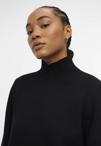 Maglione nero a collo alto con colletto a coste, texture morbida e vestibilità rilassata, indossato da un modello con capelli intrecciati, su uno sfondo neutro.