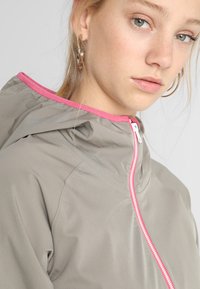 Lichte beige jas met een roze voering in de capuchon en rits. Heeft een soepele textuur en een getailleerd ontwerp.