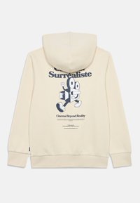 Η κρεμ-χρωματική φούτερ έχει ένα γραφικό εκτύπωμα ενός καρτούν χαρακτήρα με μεγάλα μάτια. Το κείμενο "Surrealiste" βρίσκεται πάνω και το "Cinema Beyond Reality" από κάτω.