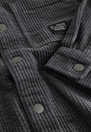 Siva kordurovina z izrazitimi navpičnimi črtami, gladkimi črnimi kovinskimi gumbi in etiketo, ki navaja velikost ter koordinate.