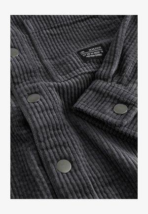 Pilkas aksominis audinys pasižymi ryškiomis vertikaliomis juostomis, lygiu juodu metaliniu mygtuku ir žyme, nurodančia dydį bei koordinates.