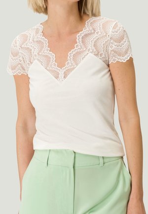 Vrouw draagt een witte top met kanten kapmouwtjes en een gegolfde V-hals, gecombineerd met lichtgroene broek met hoge taille.