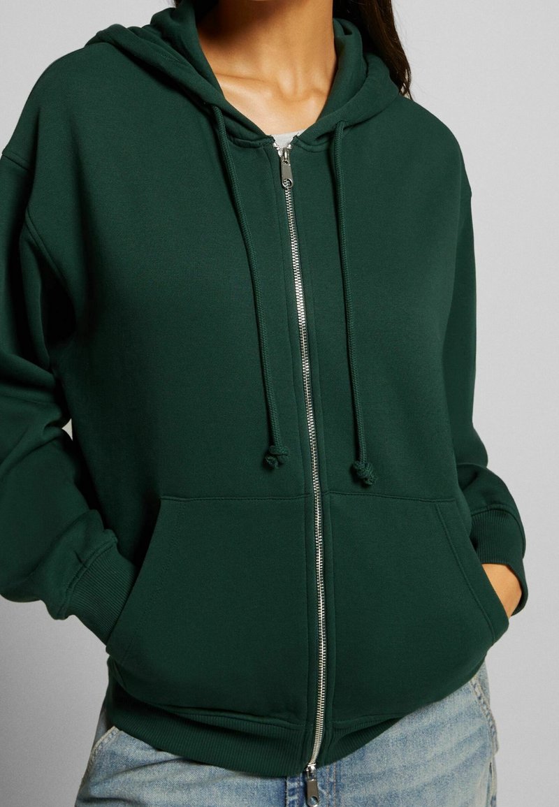 Femme portant un sweat à capuche zippé vert foncé avec cordons et poches avant, associée à un jean bleu clair, les mains dans les poches.