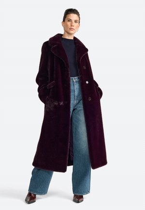 Cappotto in pelliccia sintetica lunghissimo e di un profondo colore bordeaux, con colletto a punte, bottoni frontali e tasche laterali, abbinato a jeans in denim a gamba larga e stivali.