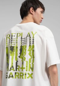 Biele oversized tričko s veľkým zeleno-čiernym grafickým potlačou slov "REPLAY MARTIN GARRIX" na chrbte v rôznych textúrach.