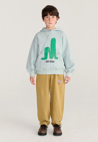 Garçon portant un sweat à capuche bleu clair avec un monstre vert de dessin animé et un pantalon moutarde avec un petit patch coloré, debout devant un fond uni.