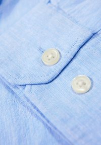 Lichtblauwe stof met een gestructureerde weving, voorzien van twee ronde, lichtgekleurde knopen en een opgevouwen kraagdetail.
