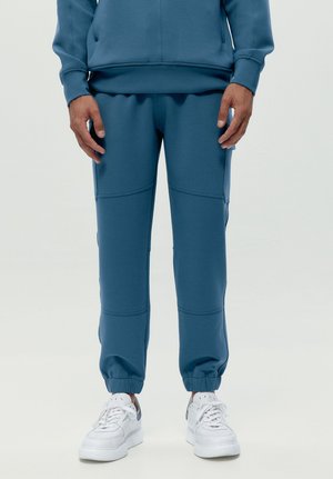 Homme portant un pantalon de jogging bleu avec des poignets élastiques et des baskets blanches, debout devant un fond clair uni.