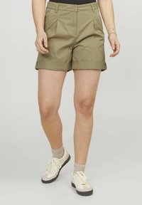 Pantaloni corti color khaki con una vestibilità rilassata, orlo risvolto, pieghe frontali e tasche laterali; abbinati a sneaker bianche e beige.