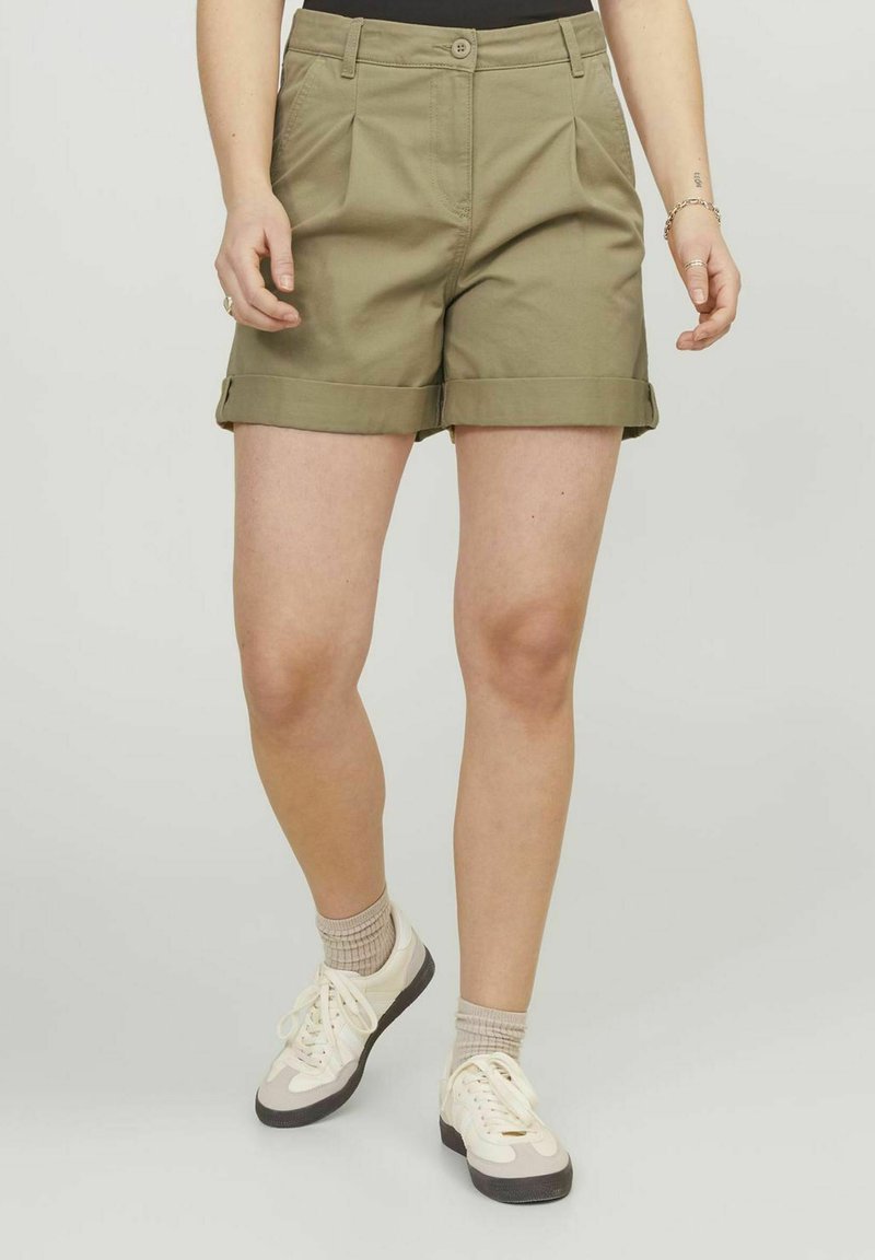 Pantaloni corti color khaki con una vestibilità rilassata, orlo risvolto, pieghe frontali e tasche laterali; abbinati a sneaker bianche e beige.