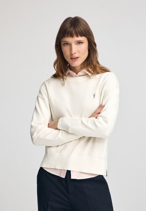 Suéter blanco con cuello redondo y puños acanalados, superpuesto sobre una camisa de cuadros rosa claro, que presenta un pequeño logotipo bordado en el pecho.