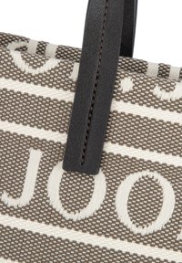 Zbliżenie na torbę z plecionego materiału z napisem "J'ADIOR" w beżowym i brązowym kolorze, z przyszytym czarnym skórzanym uchwytem.