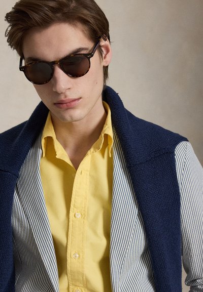 Jeune homme portant des lunettes de soleil écaille, une chemise jaune, un blazer rayé et un pull bleu marine posé sur les épaules, sur un fond uni.
