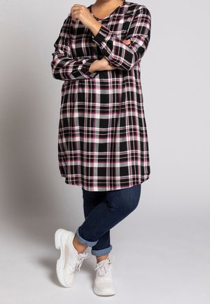 Kvinde i sort, hvid og lyserød ternet kjole kombineret med mørke opklemte jeans og hvide sneakers, stående med armene krydset.