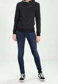 Svart tröja med vita prickar, halsringning med cowl, tre knappar och ribbade mudd. Mörkblå tajta jeans och svarta sneakers.