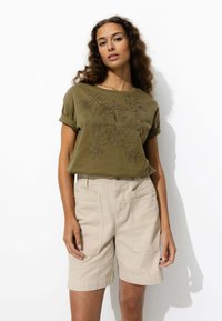 Chemise à manches courtes vert olive avec un motif floral brodé ; associée à un short beige avec des poches latérales et des ourlets retroussés.