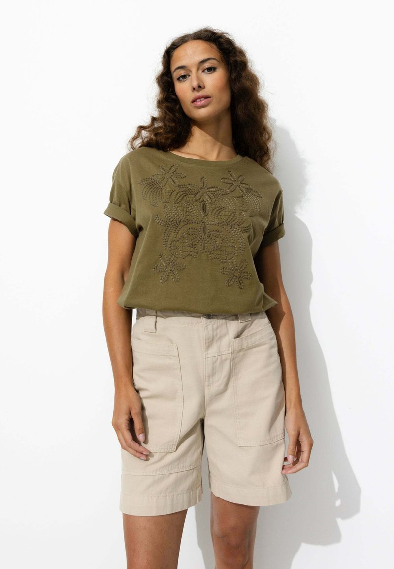 Chemise à manches courtes vert olive avec un motif floral brodé ; associée à un short beige avec des poches latérales et des ourlets retroussés.