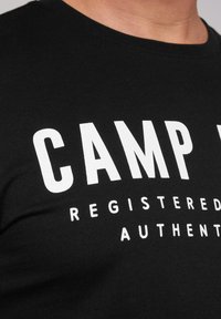 Camiseta de algodón negra con texto blanco en negrita "CAMP" y texto adicional más pequeño. La vista en primer plano enfatiza la textura y la claridad de la letra.