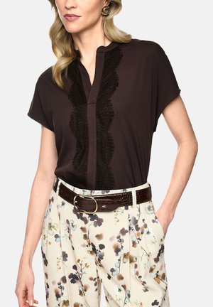 Frau trägt eine dunkelbraune Bluse mit Rüschensaum und cremefarbene, geblümte Hose, ergänzt durch einen braunen Ledergürtel mit goldener Schnalle.