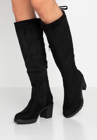 Bottes noires au-dessus du genou en suédine, avec un talon carré, une fermeture éclair latérale et des lacets en accent sur le haut, au toucher lisse.