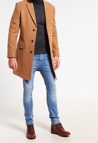 Abrigo de lana marrón claro con solapas de muesca y tres botones, llevado sobre un suéter de cuello alto oscuro. Jeans azul claro y botas de cuero marrón de tobillo complementan el look.