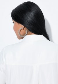 White blouse with a smooth texture, featuring a back seam and a collar. Large, circular rhinestone-studded hoop earrings.