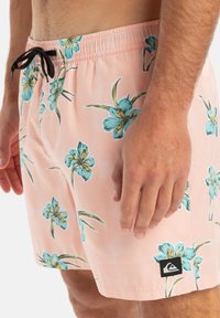 Herren-Badehosen aus leicht pinkem Stoff mit einem floralen Muster aus blauen Blumen und grünen Blättern. Enthält einen schwarzen Kordelzug und ein Logo-Patch.