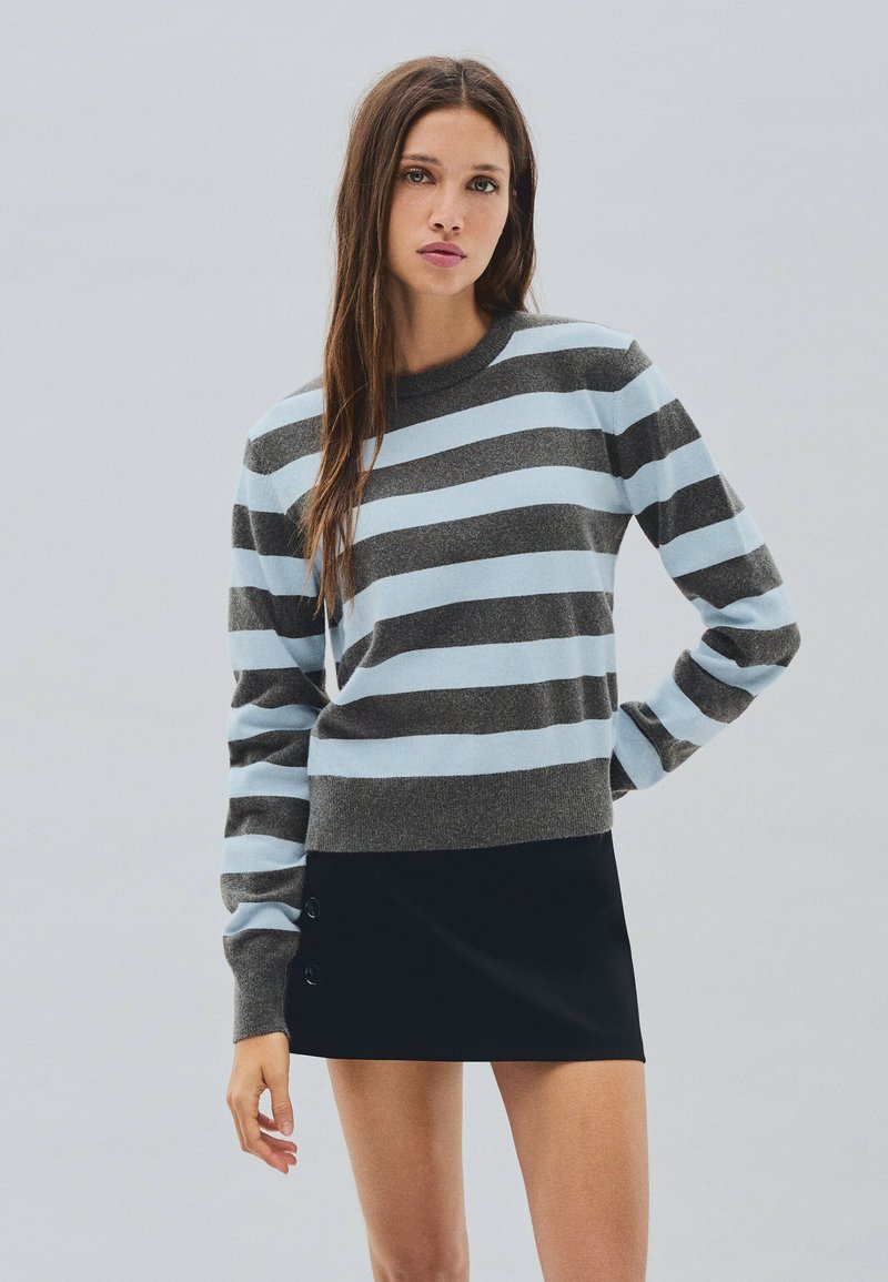 Femme aux longs cheveux bruns vêtue d'un pull rayé gris et bleu clair et d'une jupe noire courte sur un fond gris uni.