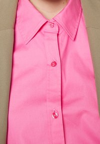 Camicia button-up rosa brillante con colletto, caratterizzata da bottoni lucidi. Un blazer beige è parzialmente visibile sul lato sinistro.