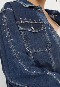 Džínová bunda v tmavě modré barvě se stříbrnými cvočky podél rukávů a na kapsách. Má zapínání na knoflíky a klasický límečkový design.