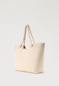 Τσάντα ώμου από μπεζ καμβά με χοντρές στριμμένες λαβές από σχοινί και ραμμένη επένδυση μπροστά σε απλό λευκό φόντο.