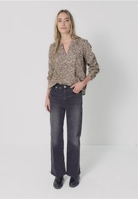 Kvinna som står klädd i en beige leopardmönstrad blus, svarta högmidjade jeans och svarta skor mot en enkel ljus bakgrund.