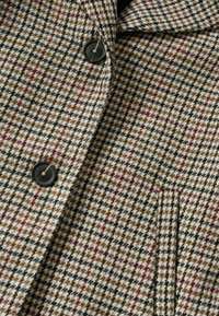 Villatakki, jossa on houndstooth-kuvio valkoisella, harmaalla ja ruskealla sävyllä. Varustettu mustilla napeilla ja yhdellä welt-taskulla.