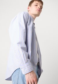 Γαλάζιο πουκάμισο με κουμπιά και κατακόρυφες ρίγες σε ανοιχτό κίτρινο. Μακριά μανίκια με κουμπωμένα μανίκια. Μαλακό ύφασμα, άνετη εφαρμογή και καμπυλωτό τελείωμα.