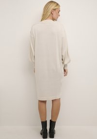 Hellbeige Oversized-Sweaterkleid, knielang, weiche Textur, gerippte Bündchen und abgerundeter Saum. Kombiniert mit schwarzen Stiefeletten.