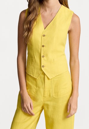 Kobieta w żółtej kamizelce bez rękawów zapinanej na guziki i pasujących do niej spódnicach z wysokim stanem na jednolitym tle.