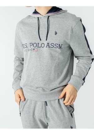 Osoba ubrana w szarą bluzę z kapturem U.S. Polo Assn. oraz pasujące spodnie dresowe z rękami w kieszeniach, pokazująca przednie i boczne paski.