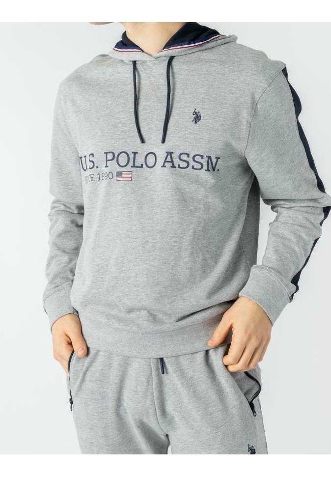 Personne portant un sweat à capuche gris U.S. Polo Assn. et un pantalon de survêtement assorti, les mains dans les poches, montrant les détails des bandes à l'avant et sur les côtés.