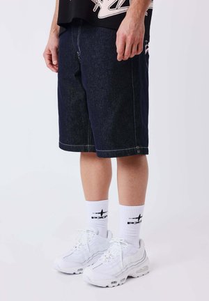 Man draagt donkere denim shorts tot op de knie, witte sokken met zwart logo en witte sportschoenen, staand tegen een effen achtergrond.
