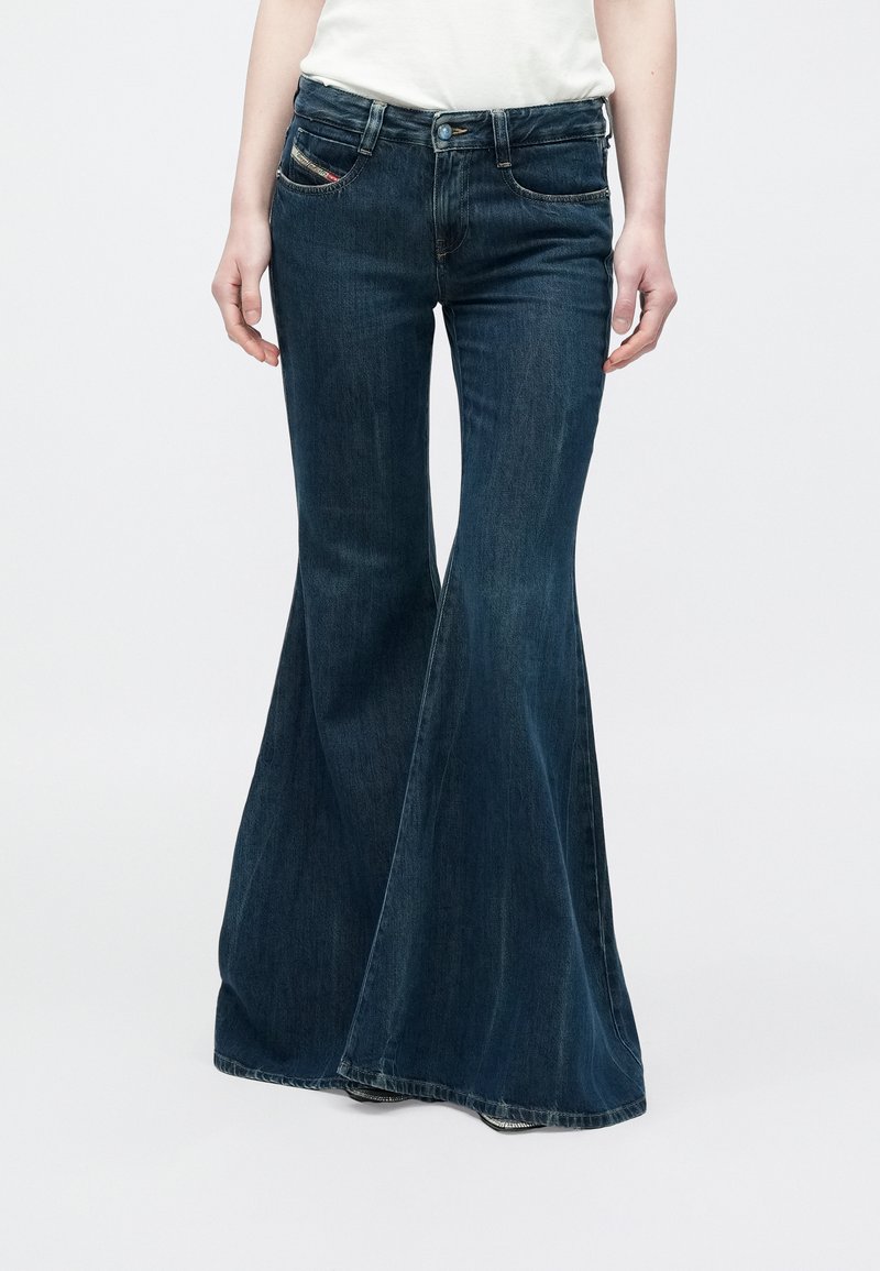 Osoba oblečená v tmavomodrých širokých džínsoch a bielom topu stojaca pred sivým jednoduchým pozadím.