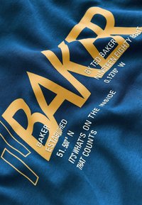 Blauwe stof met een zachte textuur toont gedurfde gouden letters, waaronder "BAKER" en "BY TED BAKER," samen met coördinaten en een slogan.