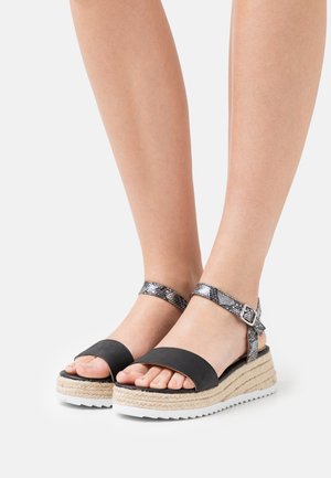 Pieds portant des sandales à plateforme noires avec des semelles en jute et des brides de cheville à motif peau de serpent sur fond blanc.