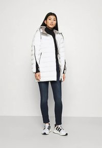 Γυναίκα που στέκεται φορώντας ένα άσπρο μπουφάν, μαύρο πουλόβερ, σκούρα τζιν και λευκά sneakers με μαύρες λεπτομέρειες, σε έναν απλό φόντο.