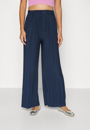 Kobieta ubrana w granatowe szerokie spodnie plisowane, srebrne baleriny i krótki różowy top, stojąca na tle jednolitego tła.